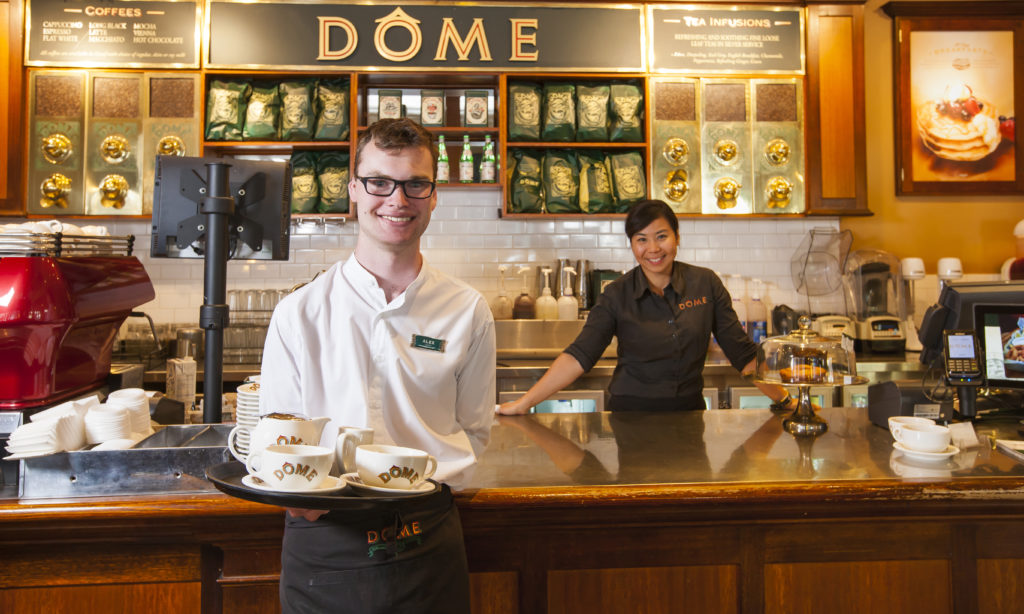Alexander holding a tray of coffees in front of the counter, while his manager stands behind the counter