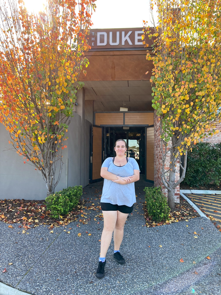 Belinda smiling outside of a building with a sign that reads The Duke