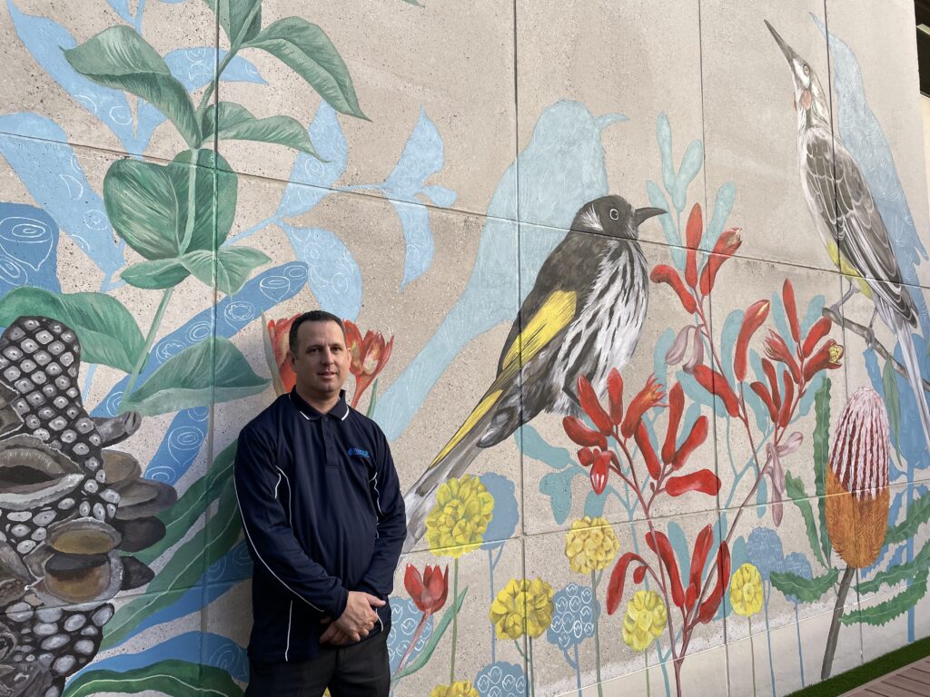 Shane smiling in front of an outdoor wall with colourful painted art.