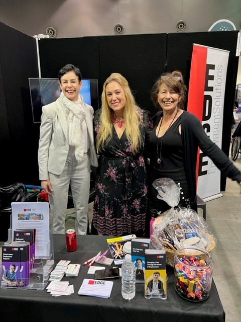 Three women standing together smiling behind a table at a expo.