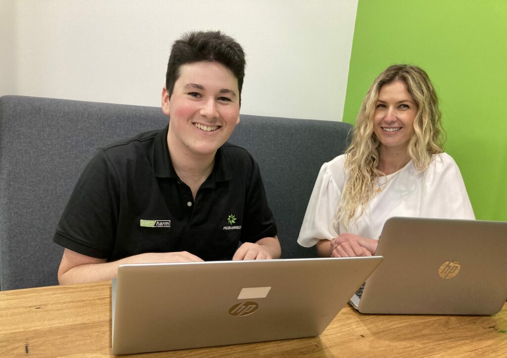 Max and Brooke sitting at a table, both with their laptops opened in front of them and smiling.