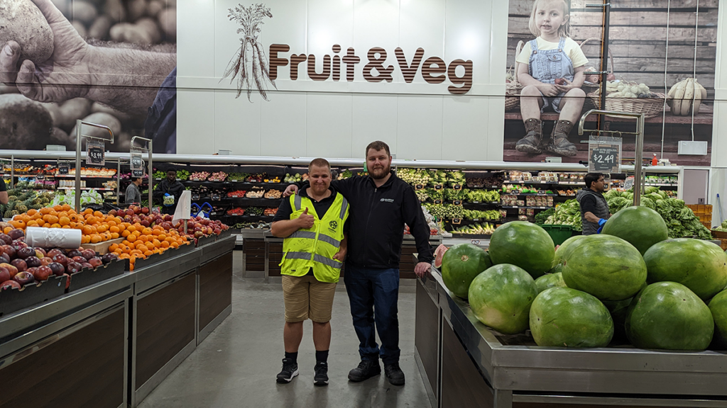 Samuel and Rhory standing together under the Fruit & Veg sign in the fruit isle at Spudshed.