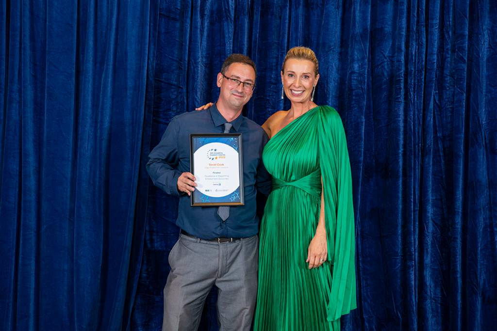 David standing next to a woman in a green dress who is also smiling. David his holding a certificate.