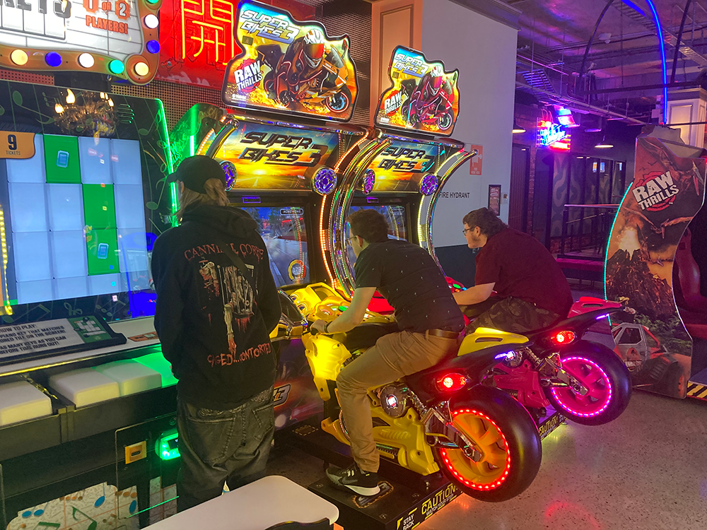 Two students sitting on motorbikes while another stands playing an arcade game at Strike Bowling.