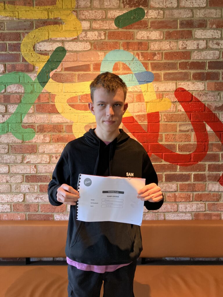 Daniel holding a piece of paper in front of a brick wall at the Grill'd store he works at.
