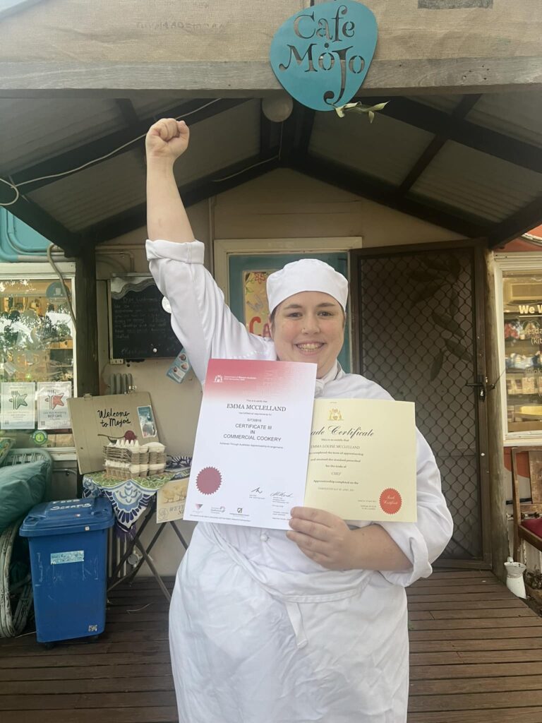 Emma holding two certificates in Chef's uniform while reaching one hand up above her head.