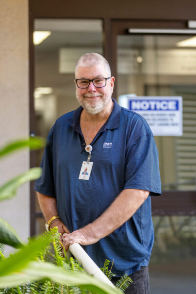 A mid shot of Philip smiling in front of the hospital door while he rests his hands on a hand rail.