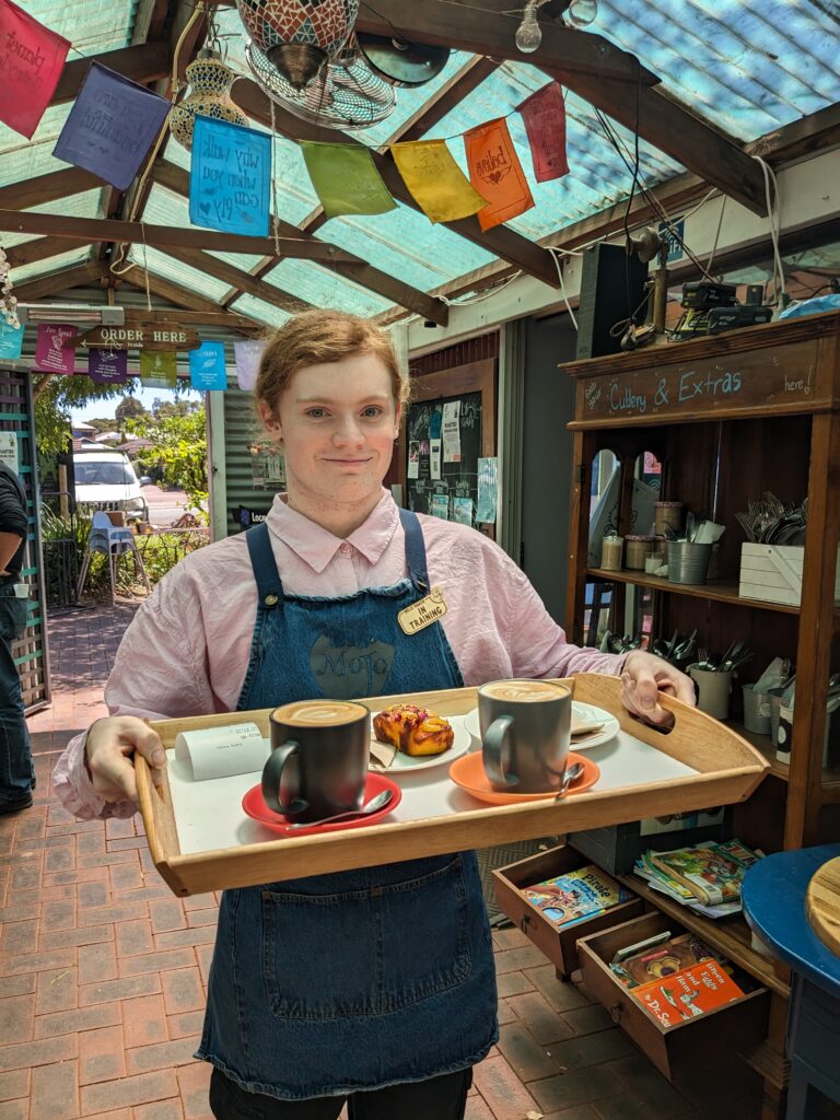 Image of Zoe holding a wooden tray with two coffee cups and food in it. Zoe is wearing a pink shirt and apron.
