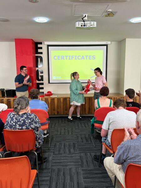 A group of people seated on red chairs facing two staff members presenting a student with a graduation folder in front of projector with a yellow background and the word Certificates.
