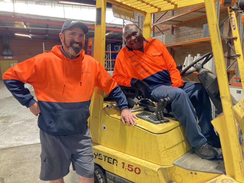 Two men in a warehouse with one sitting on a forklift while the other man is standing leaning against the yellow forklift.