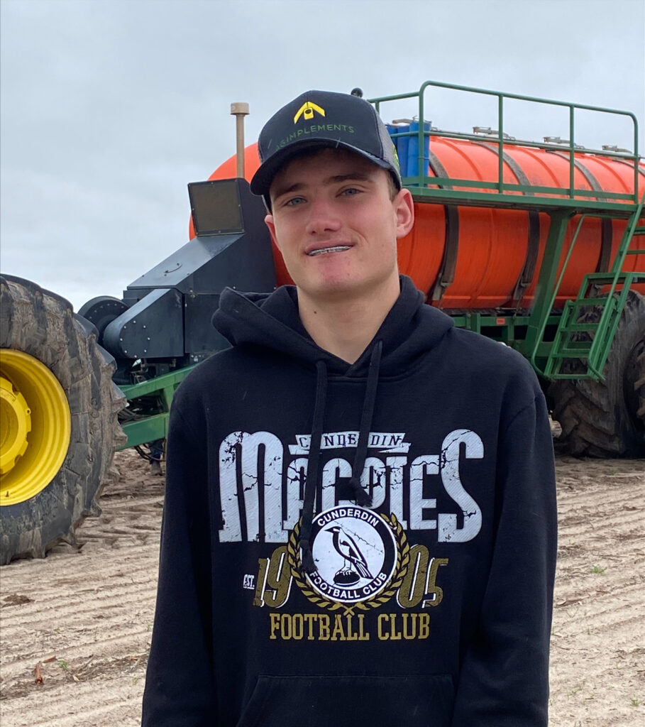 Jack wearing a jumper with graphics and a hat standing in front of a seeder box.