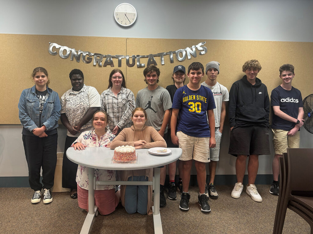 A group of students standing around a small table with a cake. Behind them is a sign hanging up with the word, congratulations.