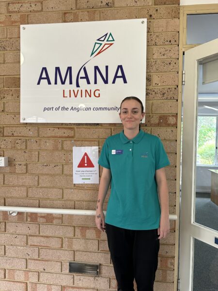 Jasmine standing in front of a brick wall with a white sign of the Amana Living logo.