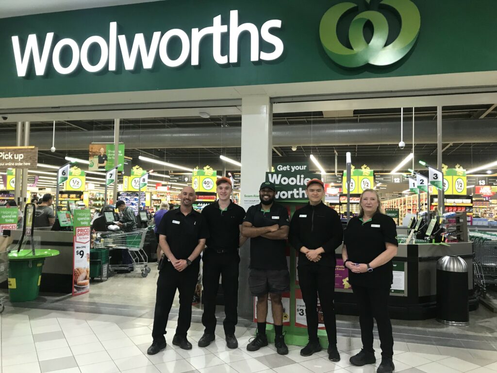 Five people standing side-by-side while all smiling at the front of a Woolworths store. Above them is a Woolworths sign.