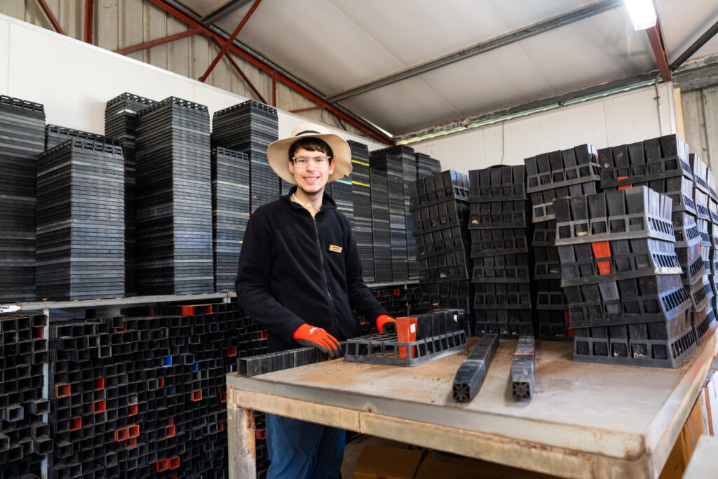 A smiling young man wearing glasses, a wide-brimmed hat, red work gloves, and a dark jacket stands at a workbench inside a warehouse. He is handling black plastic seedling trays, with large stacks of similar trays surrounding him on shelves and pallets. The space has a metal roof and industrial setting.