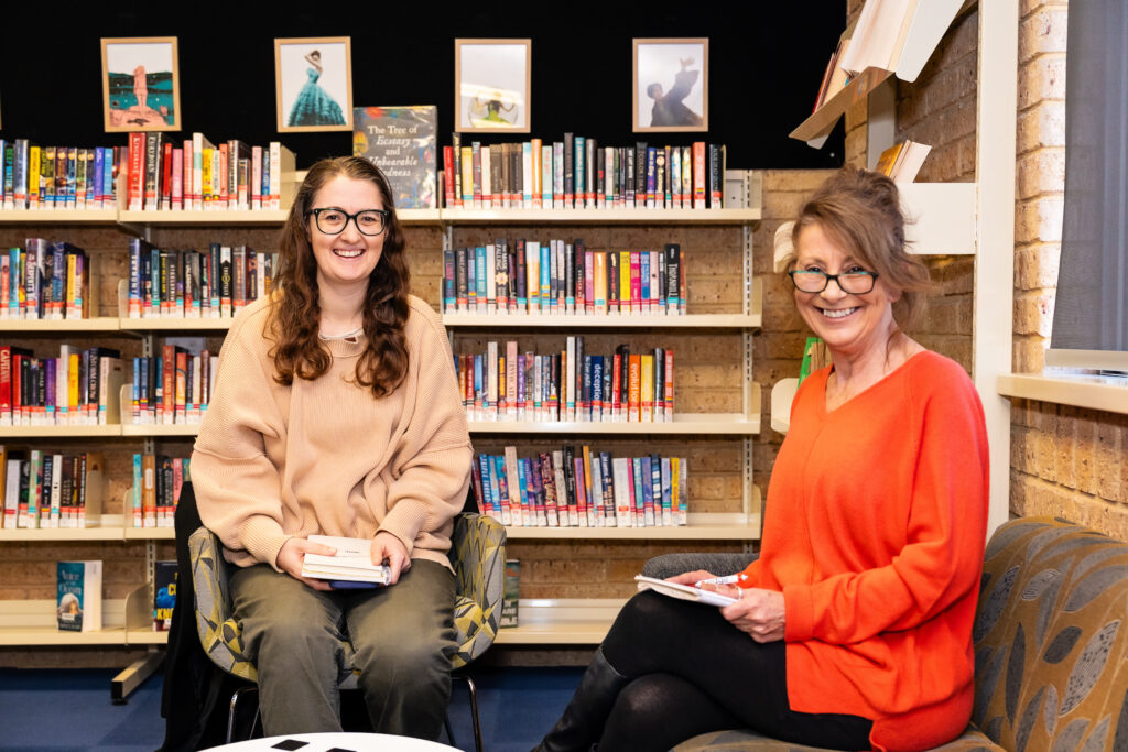 Two woman sitting side by side on chairs inside a library.