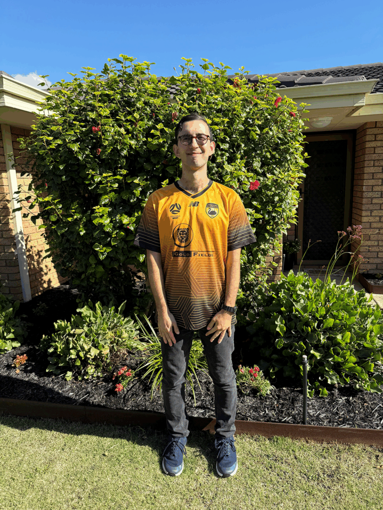 Full body image of Luka standing outside smiling. He is wearing a gold Western Australian state football shirt.