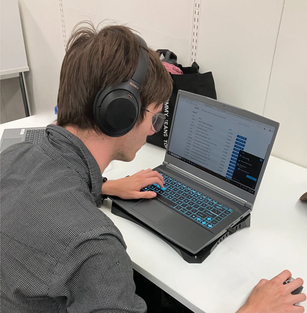 A young man wearing over-ear headphones works on a laptop at a white desk. The laptop sits on a stand with a backlit keyboard glowing blue. He uses a mouse with his right hand while looking at a screen displaying a list-style interface. In the bottom left corner, the Edge logo appears with the tagline 'Beyond work. Beyond disability.'