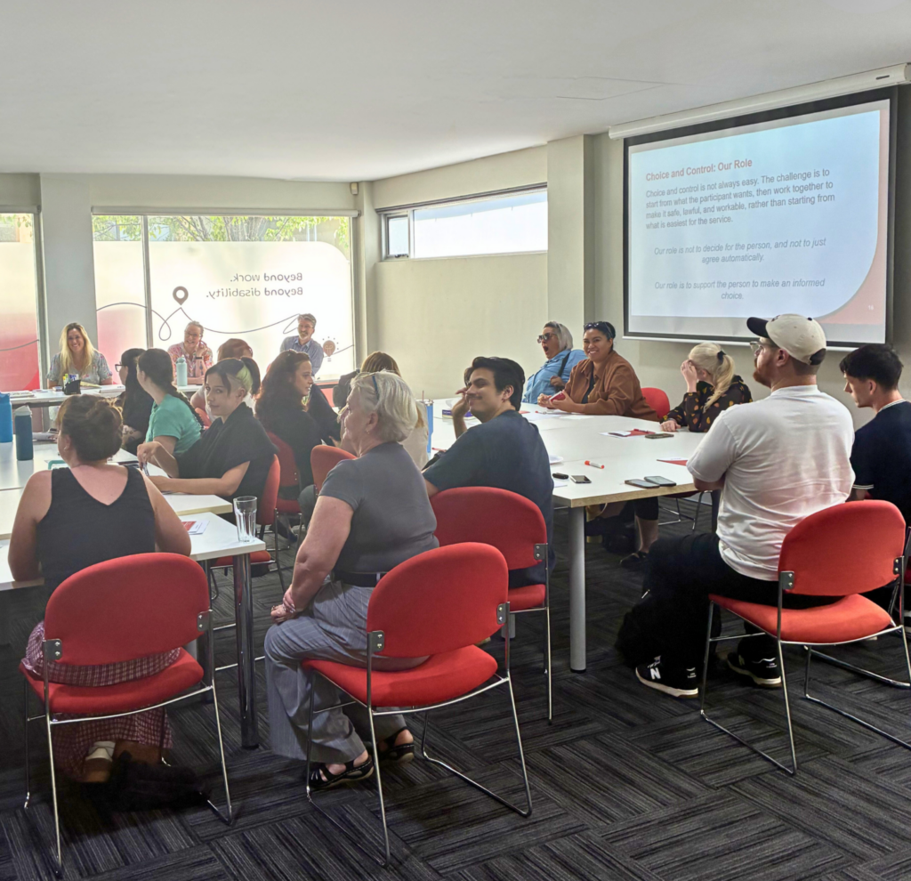 A group of staff seated at tables in a training room during a professional development session. Staff are seated on red chairs around white tables, listening and smiling as a presentation is shown on a screen at the front of the room. The slide reads “Choice and Control: Our Role.” The image is framed with a red graphic border.