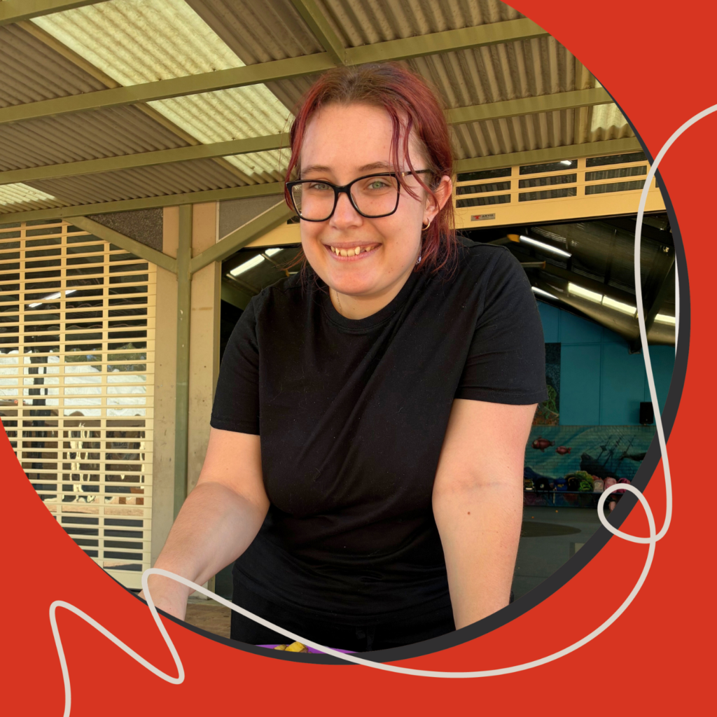 A smiling person with red hair and glasses leans forward over a table, preparing food outdoors under a covered structure. The woman is wearing a black shirt and holding a purple plate with chopped ingredients.