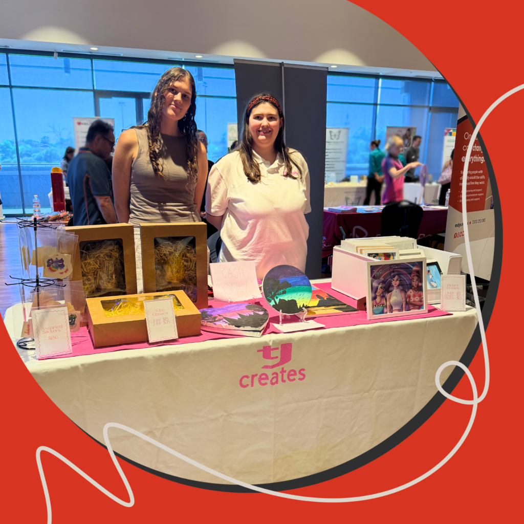 Two women stand behind a market stall at a disability expo, smiling at the camera. The table, branded “tj creates,” displays handmade items. The image is presented within a circular frame on a red graphic background, with other expo stalls and attendees visible behind them.
