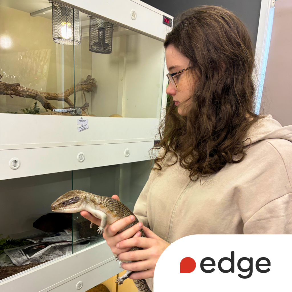 A young woman with glasses holds a lizard while standing beside animal enclosures at a wildlife hospital. She is wearing a beige hoodie and looking at the lizard. The Edge logo appears in the bottom corner of the image.