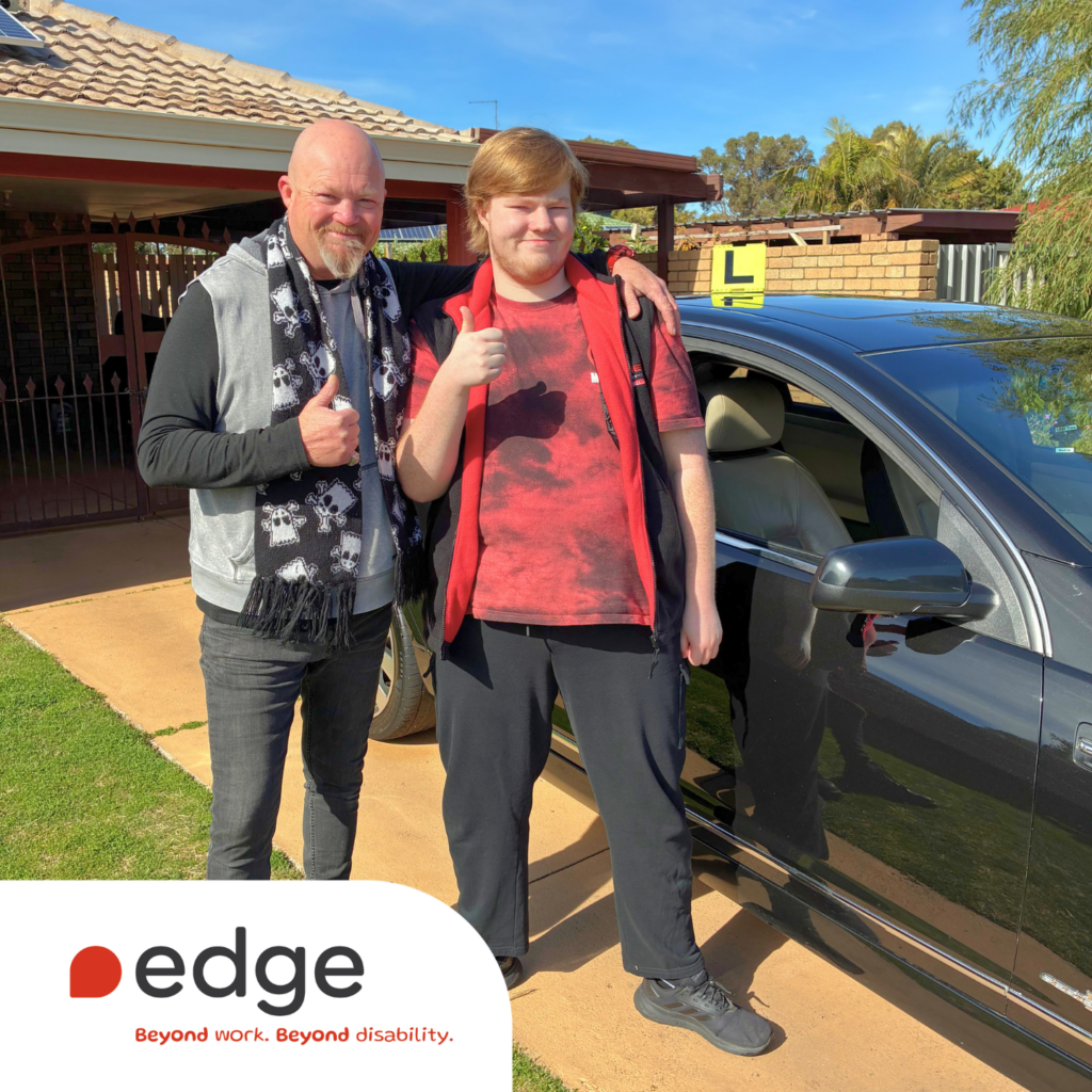 A smiling young learner driver stands beside a black car with an L-plate on the roof, giving a thumbs up and is standing next to a man, who is also giving a thumbs up. They are standing in a sunny residential driveway. The Edge logo appears in the bottom left corner of the image.