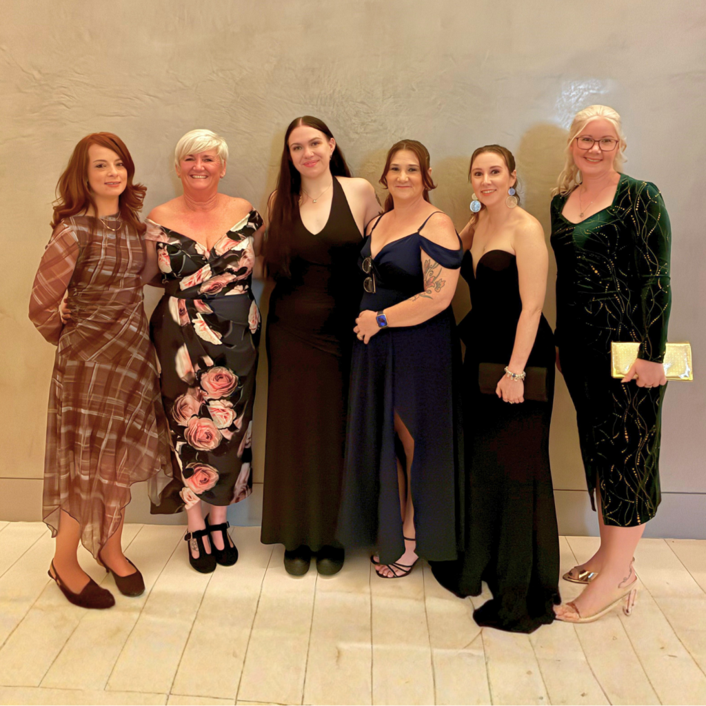 Six women dressed in elegant evening gowns stand side by side against a neutral textured wall, smiling at the camera. Their dresses range from patterned and floral styles to sleek black formal wear, with accessories like earrings, clutches, and heels. In the lower corner, our logo reads 'Edge – Beyond work. Beyond disability.'