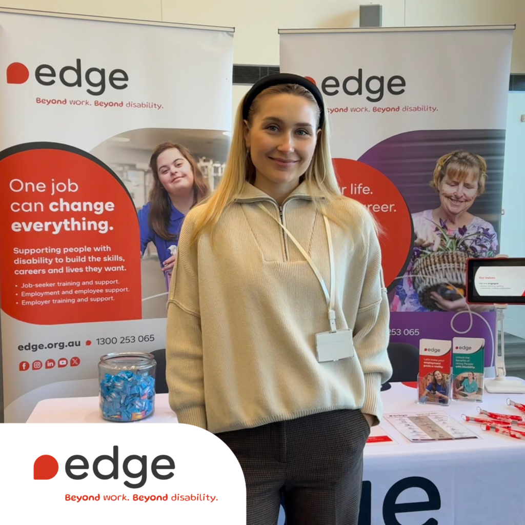 A smiling Edge team member stands in front of an Edge booth at an Expo. Behind her are two Edge banners promoting employment support for people with disability, along with brochures, pens, and a jar of mints on the table. The Edge logo and tagline "Beyond work. Beyond disability." appear in the bottom left corner of the image.