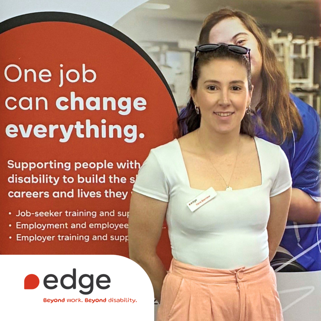 A mid shot of Tara smiling. She is wearing a white top and peach trousers and stands in front of an Edge banner. The banner reads, "One job can change everything", and highlights support for people with disability to build careers and lives, alongside the Edge logo and the tagline, 'Beyond work. Beyond disability.'