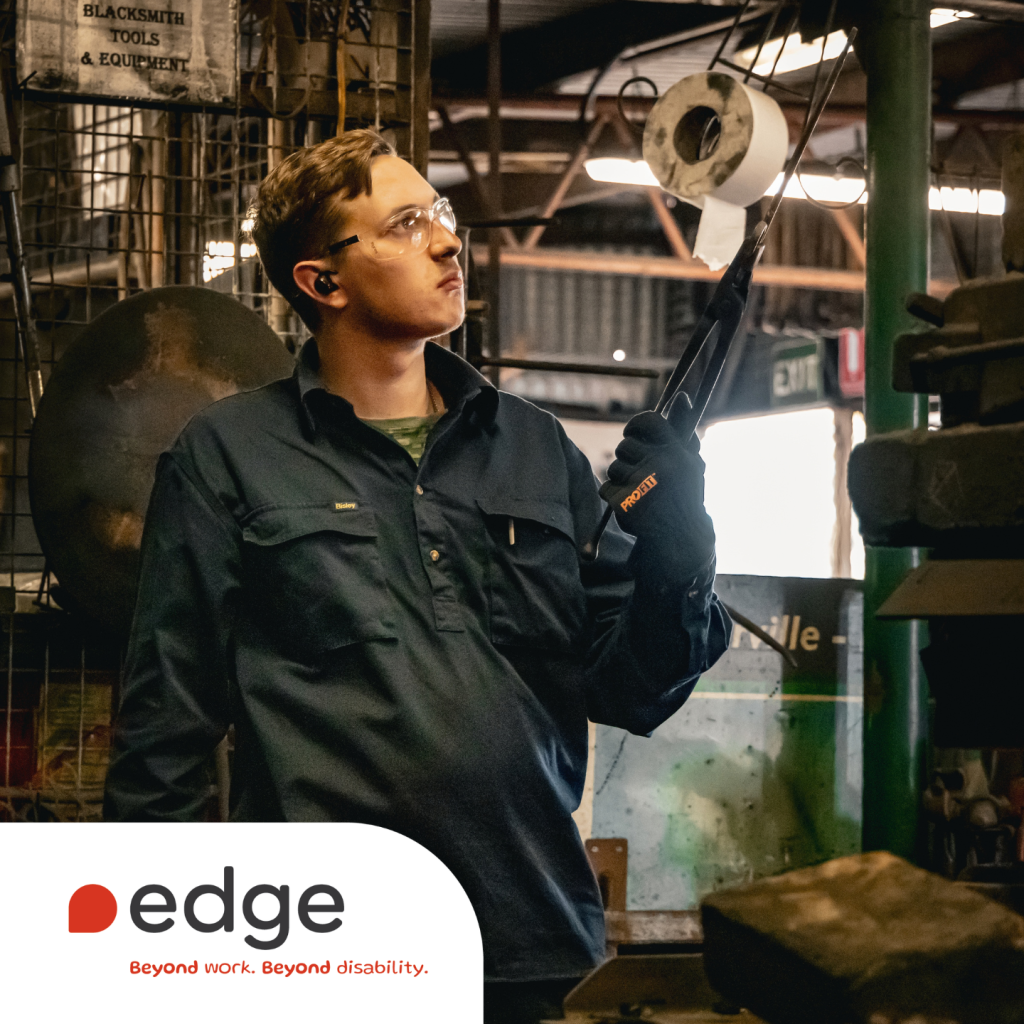 Ash wearing protective eyewear, gloves and workwear while holding blacksmithing tools in a workshop. Edge Employment Solutions branding and the tagline “Beyond work. Beyond disability.” appear at the bottom of the image.